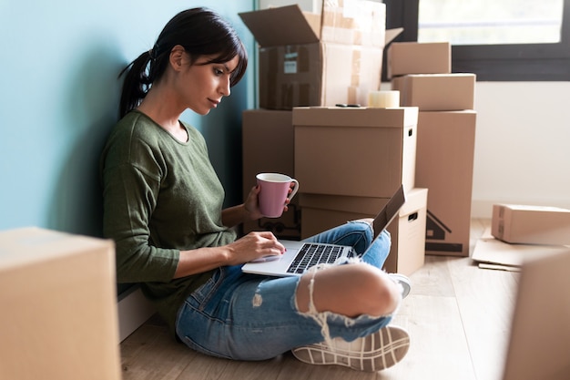 Aufnahme einer schönen jungen Frau, die mit Laptop arbeitet, während sie zu Hause auf dem Boden sitzt und Kaffee trinkt.