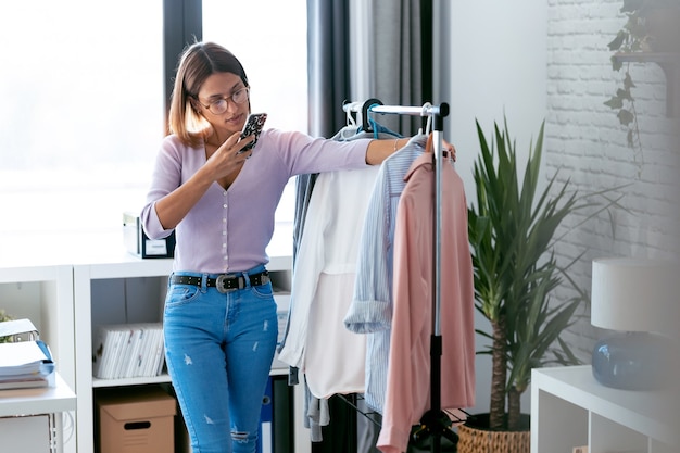Aufnahme einer schönen jungen Frau, die Outfit sucht und auswählt, während sie Fotos mit ihrem Smartphone in der heimischen Garderobe macht.