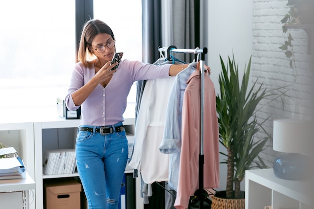 Aufnahme einer schönen jungen Frau, die Outfit sucht und auswählt, während sie Fotos mit ihrem Smartphone in der heimischen Garderobe macht.