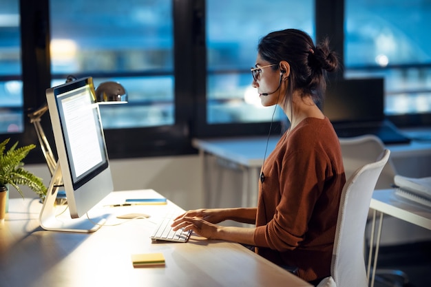 Aufnahme einer schönen jungen Geschäftsfrau, die mit Computer arbeitet, während sie mit Kopfhörer im Büro spricht.