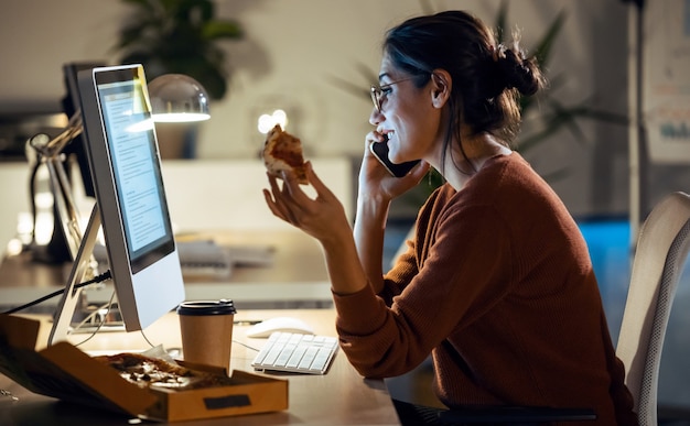 Aufnahme einer schönen jungen Geschäftsfrau, die mit Computer arbeitet, während sie Pizza im Büro isst.