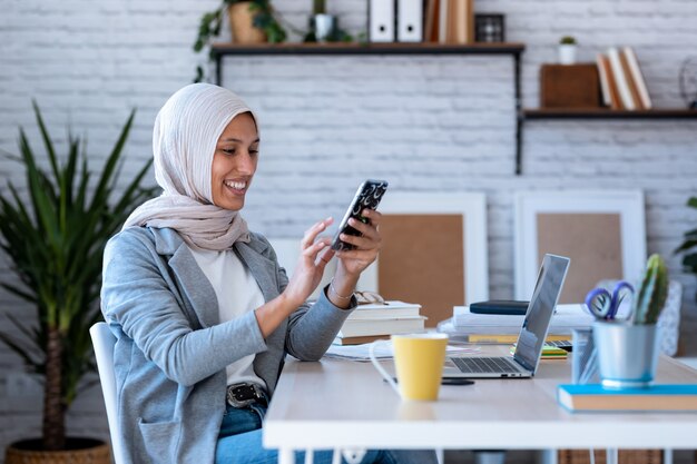 Aufnahme einer schönen jungen muslimischen Geschäftsfrau mit Hijab, die Nachrichten mit dem Handy sendet, während sie im Büro sitzt.