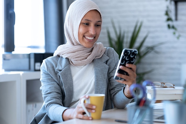 Aufnahme einer schönen jungen muslimischen Geschäftsfrau mit Hijab, die Nachrichten mit dem Handy sendet, während sie im Büro sitzt.