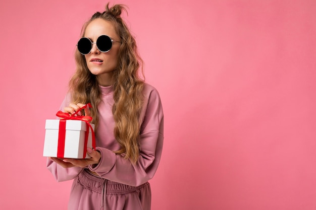 Aufnahme einer schönen positiven jungen blonden lockigen Frau, die über rosafarbener Hintergrundwand isoliert ist und rosa Sportkleidung und Sonnenbrille trägt, die Geschenkbox hält und Überraschung auspackt, die zur Seite schaut. Freiraum