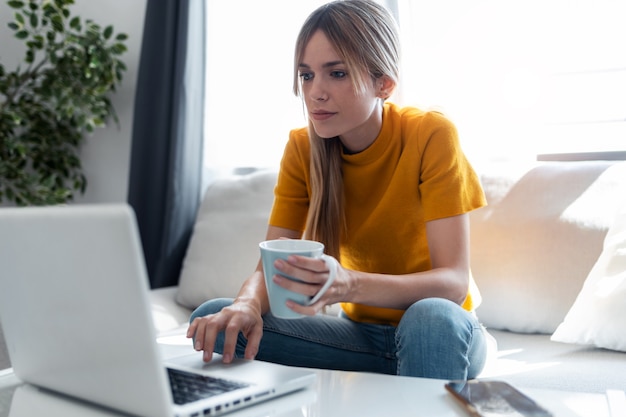 Aufnahme einer selbstbewussten jungen Frau, die mit ihrem Laptop arbeitet, während sie eine Tasse Kaffee trinkt, die zu Hause auf einer Couch sitzt