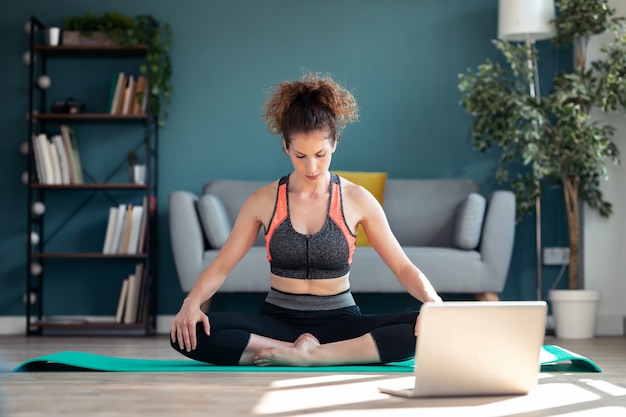 Aufnahme einer selbstbewussten, sportlichen jungen frau, die nach online-fitness-kursen über laptop auf dem boden in ihrem wohnzimmer zu hause hypopressive übungen macht.