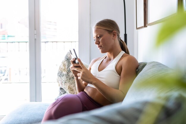 Aufnahme einer sportlichen glücklichen Frau, die Nachrichten mit ihrem Smartphone sendet, während sie zu Hause auf der Couch im Wohnzimmer sitzt.