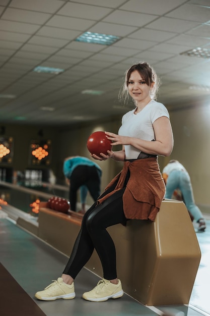 Aufnahme einer süßen jungen Frau, die eine Bowlingkugel im Bowlingclub hält und in die Kamera schaut.