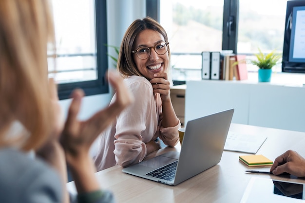 Aufnahme einer süßen jungen Frau, die ihrem Kollegen zuhört und lächelt, während sie mit dem Computer am Coworking-Platz arbeitet.