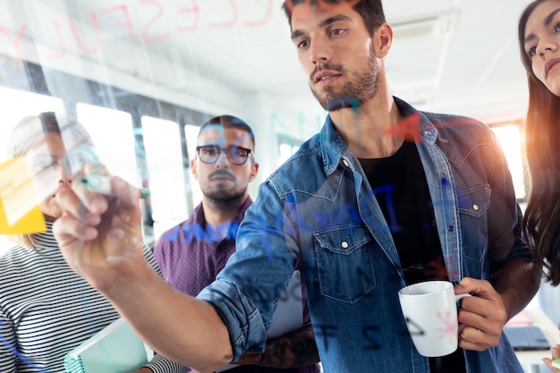 Aufnahme eines erfolgreichen jungen Mannes, der während des Treffens mit Kollegen im Coworking Space auf einer Büroglastafel schreibt.