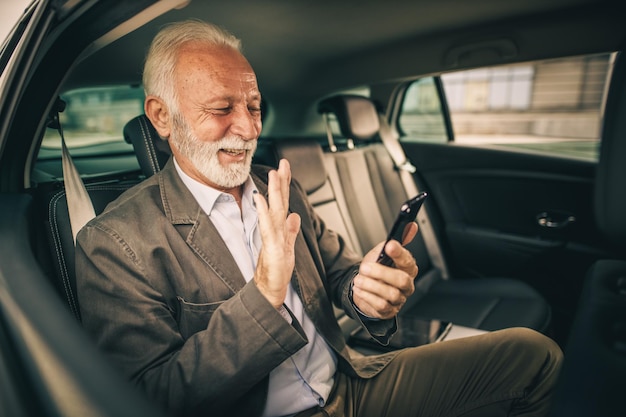 Aufnahme eines erfolgreichen Senior-Geschäftsmanns, der einen Videoanruf auf einem Smartphone tätigt, während er während seiner morgendlichen Fahrt zur Arbeit auf dem Rücksitz eines Autos sitzt.