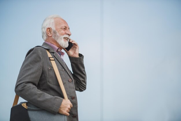 Aufnahme eines erfolgreichen Senior-Geschäftsmanns, der vor einem Firmengebäude mit einem Smartphone spricht.