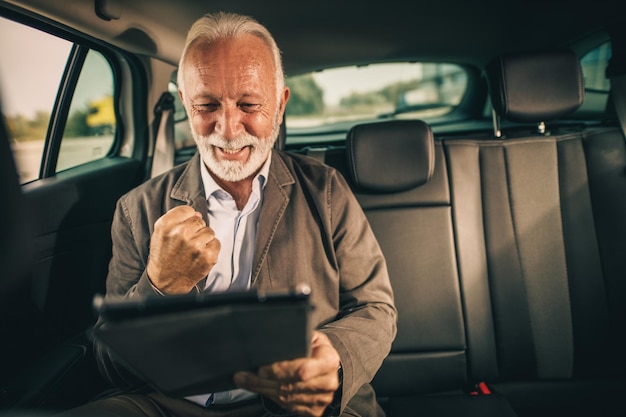 Aufnahme eines erfolgreichen Senior-Geschäftsmanns, der während seiner morgendlichen Fahrt zur Arbeit auf dem Rücksitz eines Autos sitzt und ein digitales Tablet verwendet.