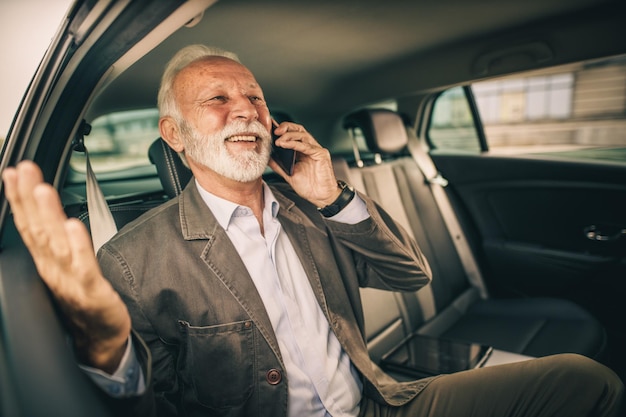 Aufnahme eines erfolgreichen Seniors, der während seiner Geschäftsreise auf dem Rücksitz eines Autos mit einem Smartphone spricht.