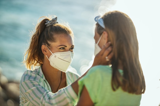 Aufnahme eines glücklichen kleinen Mädchens und ihrer jungen Mutter mit schützender N95-Maske, die bei einer Koronapandemie Zeit an der Küste verbringen.