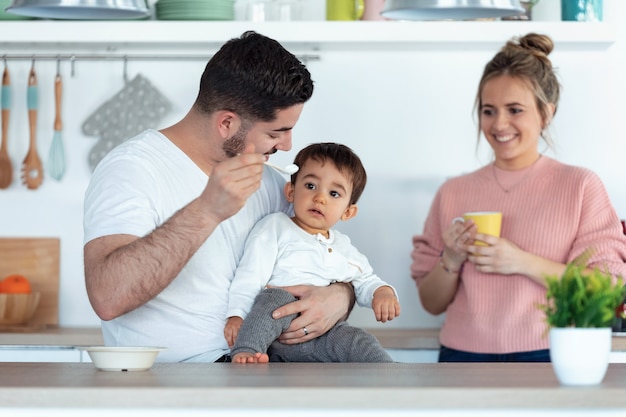 Aufnahme eines hübschen jungen Vaters, der seinen kleinen Sohn füttert, während die Mutter sie zu Hause in der Küche ansieht.