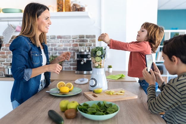 Aufnahme eines Jungen, der seiner Mutter hilft, einen Entgiftungssaft mit einem Mixer zuzubereiten, während sein Bruder in der Küche zu Hause mit dem Handy fotografiert.