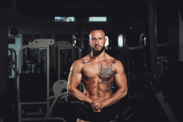 Aufnahme eines jungen muskulösen Bodybuilders, der seine perfekten Muskeln zeigt, während er nach dem Training im Fitnessstudio posiert. Blick in die Kamera.