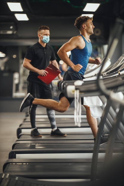 Aufnahme eines muskulösen jungen Mannes, der im Fitnessstudio auf dem Laufband läuft. Er arbeitet mit Personal Trainer.