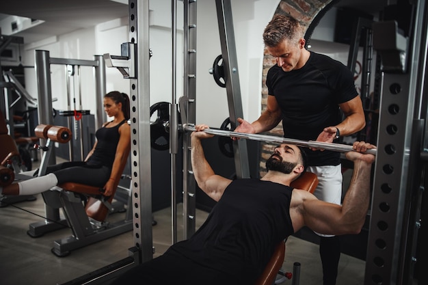 Aufnahme eines muskulösen Typen in Sportkleidung, der mit einem persönlichen Trainer im Fitnessstudio trainiert. Er pumpt die Brustmuskulatur während eines harten Trainings auf.