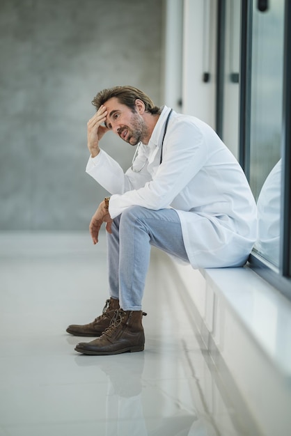 Aufnahme eines reifen Arztes, der gestresst aussieht, während er während der Covid-19-Pandemie in der Nähe eines Fensters in einem leeren Krankenhausflur sitzt.