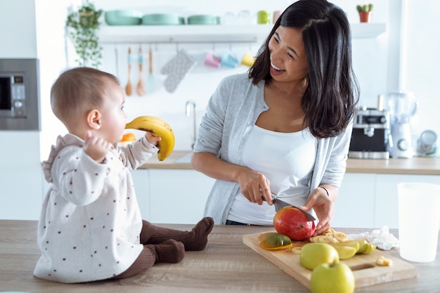 Aufnahme eines süßen kleinen Mädchens, das mit Essen spielt, während ihre Mutter Obst schneidet, um Babybrei in der Küche zuzubereiten.