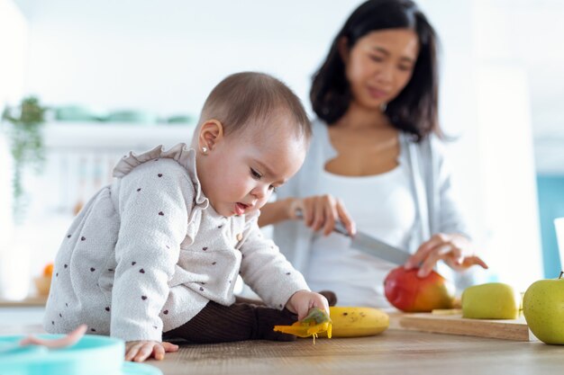 Aufnahme eines süßen kleinen Mädchens, das mit Essen spielt, während ihre Mutter Obst schneidet, um Babybrei in der Küche zuzubereiten.