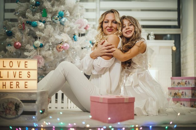 Aufnahme eines süßen kleinen Mädchens und ihrer attraktiven Mutter, die neben dem weißen Weihnachtsbaum auf dem Boden sitzen und ihre Weihnachtsgeschenke öffnen.