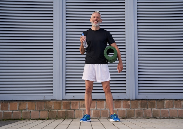 Foto aufnahme in voller länge eines aktiven sportlers mittleren alters, der eine trainingsmatte und eine flasche wasser bereithält