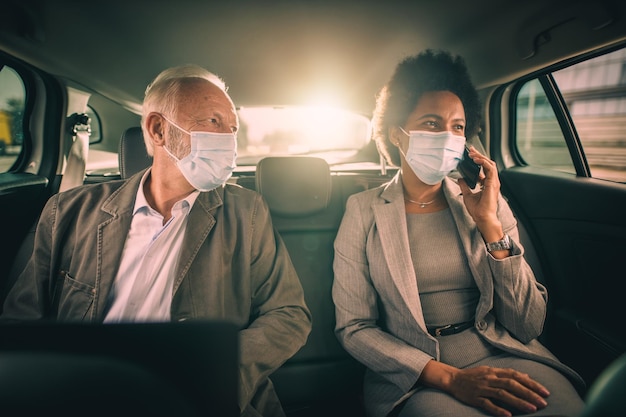 Aufnahme von zwei erfolgreichen multiethnischen Geschäftsleuten mit Schutzmaske, während sie während der COVID-19-Pandemie auf dem Rücksitz eines Autos auf ihrem morgendlichen Arbeitsweg sitzen.