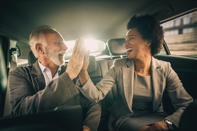 Aufnahme von zwei erfolgreichen multiethnischen Menschen, die fünf geben und gute Arbeit feiern, während sie während ihrer Geschäftsreise auf dem Rücksitz eines Autos sitzen.