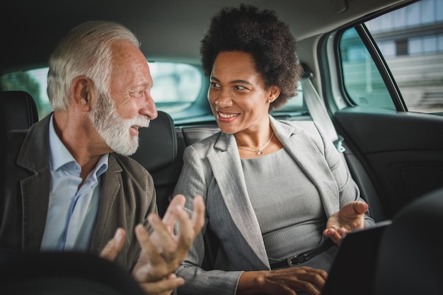 Aufnahme von zwei erfolgreichen multiethnischen Menschen, die während ihrer Geschäftsreise auf dem Rücksitz eines Autos einen Laptop benutzen und eine Diskussion führen.