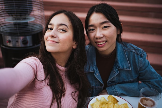 Aufnahme von zwei glücklichen Mädchen, die ein Selfie machen, während sie ein Bier und einen Snack trinken