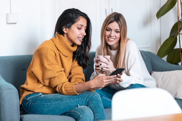 Aufnahme von zwei hübschen jungen Frauen, die Handy beim Kaffeetrinken auf dem Sofa zu Hause benutzen.