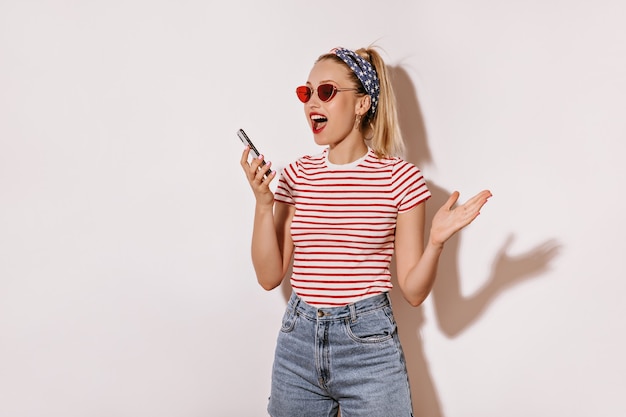 Aufregende Dame mit blonden Haaren im Stirnband, roter Sonnenbrille, gestreiftem T-Shirt und kurzem Jeans-Holding an isolierter Wand