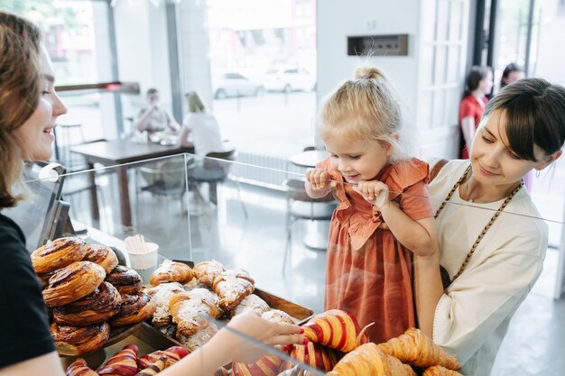 Aufregendes kleines Mädchen, das einige bunte Croissants betrachtet und auswählt, was sie will, während die Mutter sie in den Händen hält. Kassiererin lächelt sie an.