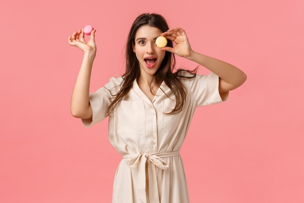 Aufregung, Urlaub und süße Desserts Konzept. Elegante attraktive junge Frau genießen köstliches Essen, Macarons auf Auge halten, offener Mund amüsiert nach Luft schnappen, stehende rosa Wand