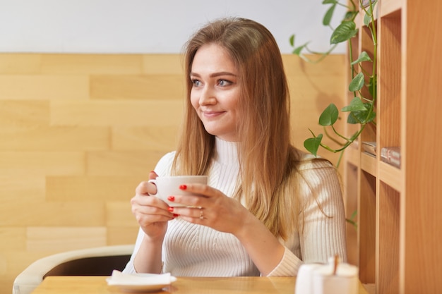 Aufrichtige süße junge Dame sitzt in der örtlichen Cafeteria in der Nähe eines Holzregals mit einer grünen Pflanze hinter sich und hält eine kleine weiße Tasse Kaffee in beiden Händen