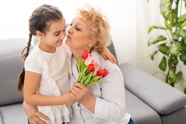 Aufrichtige, süße kleine Enkelin, die stark kuschelte und auf die Wange küsste, ihre Großmutter aus den 60er Jahren gab ihr hübsche Frühlingsblumen, die ihr zum Geburtstag, dem Internationalen Frauentag, einem Nahaufnahme-Konzeptbild gratulierten.