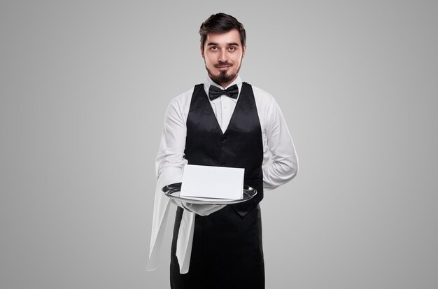 Aufrichtiger erwachsener Kellner in Uniform mit Hand hinter dem Rücken und leerem Papier auf dem Tablett mit Blick in die Kamera auf grauem Hintergrund