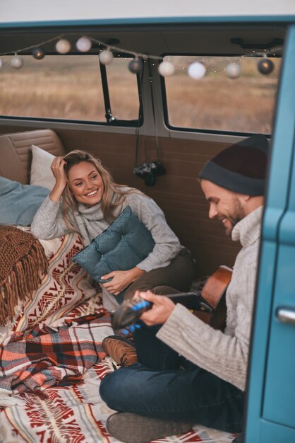 Aufrichtiges Lächeln. Draufsicht des hübschen jungen Mannes, der Gitarre für seine schöne Freundin spielt, während im blauen Retro-Art-Minivan sitzt