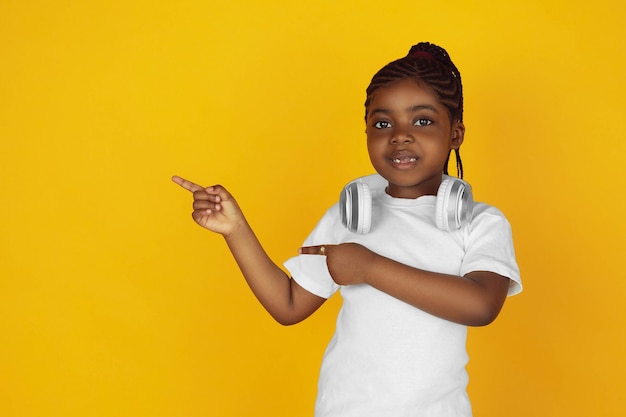 Aufzeigen, Kopfhörer aufsetzen. Porträt des kleinen afroamerikanischen Mädchens auf gelbem Studiohintergrund. Fröhliches Kind. Konzept der menschlichen Emotionen, Gesichtsausdruck, Verkauf, Anzeige. Exemplar. Sieht süß aus.