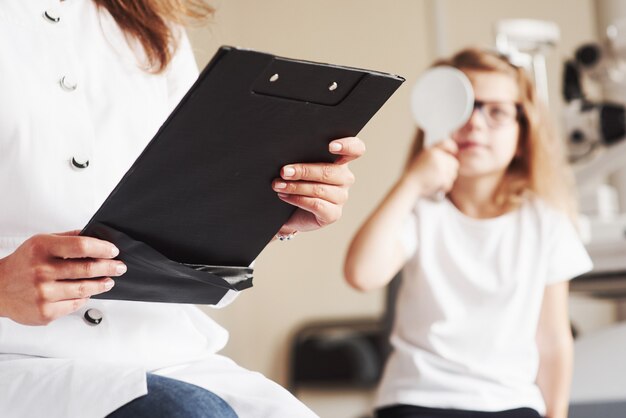 Augengesundheitskonzept. Frau mit Dokumenten testet die Sehschärfe eines kleinen Mädchens.