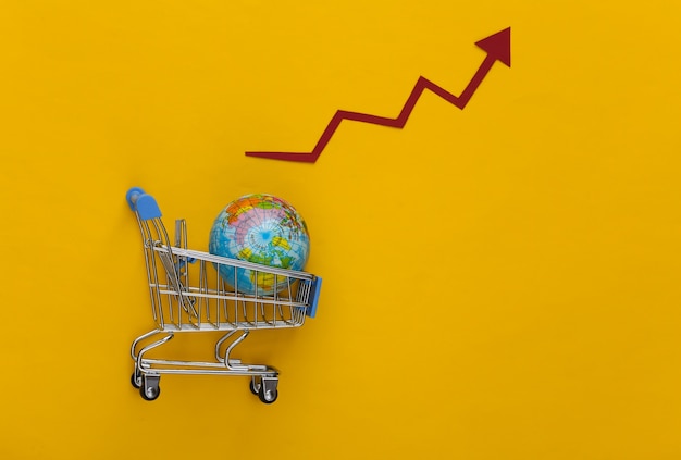 Foto aumento de las compras globales. carro de supermercado con un globo terráqueo, la flecha de crecimiento tiende hacia arriba sobre un amarillo.