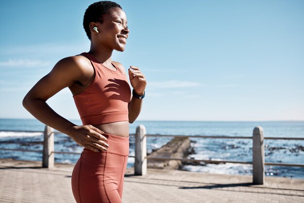 Auricular de música de mujer negra o corriendo por la playa, el océano o el mar en el bienestar de la salud o la fuerza cardiovascular Sonrisa feliz corredora deportiva o escuchando entrenamiento de entrenamiento o radio de ejercicios de maratón