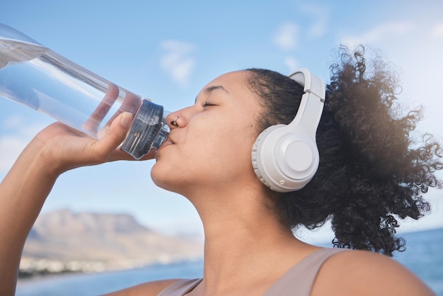 Auriculares de fitness y mujer bebiendo agua después de correr entrenamiento y ejercicio cardiovascular en la playa Corredor de ejercicio y sediento con líquido saludable para hidratar la energía y el bienestar escuchando música