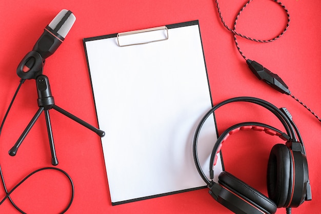 Auriculares, micrófono y portapapeles con papel blanco sobre fondo rojo. Música conceptual o podcast. Vista superior, endecha plana