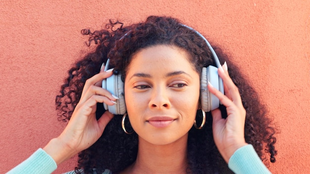 Auriculares musicales y cara de una mujer negra feliz escuchando canciones de radio y sonido contra un fondo de pared Cabeza femenina africana en emoción de felicidad inalámbrica y lista de reproducción de audio divertida al aire libre