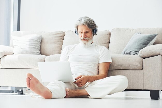 En los auriculares Se sienta cerca del sofá Hombre moderno con estilo senior con cabello gris y barba en el interior