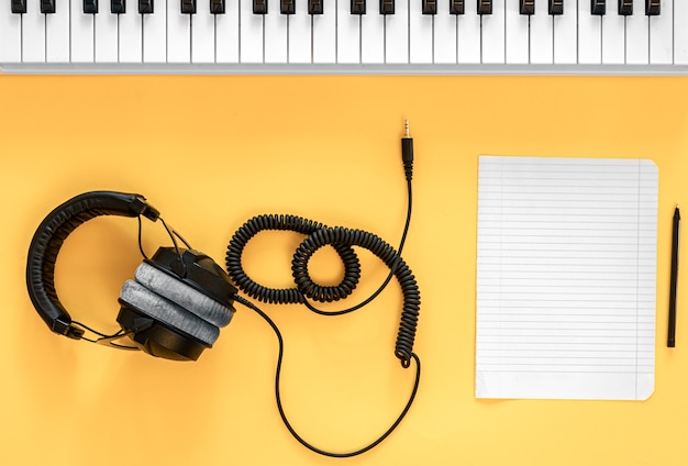 Foto auriculares, teclas musicales y una hoja de papel en blanco sobre un fondo amarillo, plano, minimalismo.